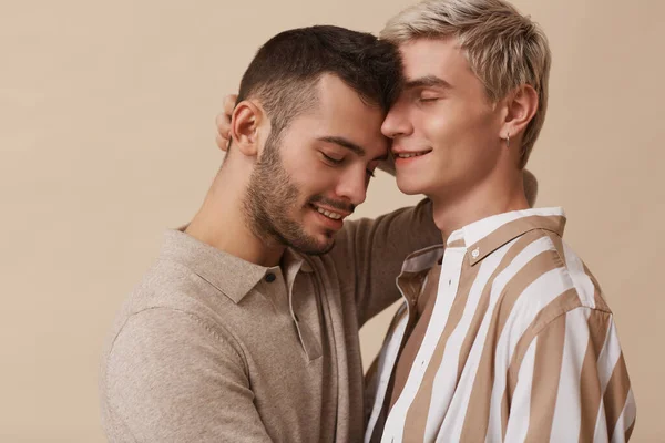 Minimal portrait of happy gay couple embracing lovingly while posing against beige background in studio