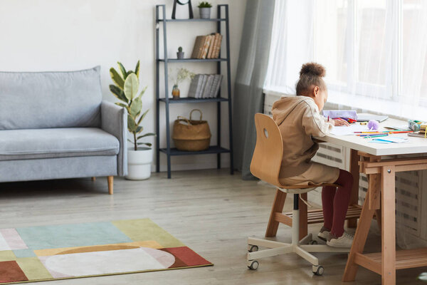 Full length portrait of cute African-American girl doing homework or drawing while sitting at desk in cozy home interior, copy space