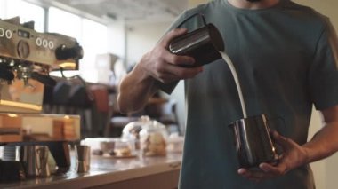 Barda otomatik kahve makinesinin yanında duran ve kapuçino için sütü sürahiden kavanoza döken tanınmamış barmenin orta boy fotoğrafı.
