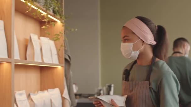 カフェで棚にコーヒーのパックを敷設制服や医療マスクを身に着けている若い混合レースの女性バーテンダーの中ショット — ストック動画