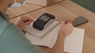 Tanımlanamayan, üniforma giyen, tıbbi maske takan, ahşap masada oturan, temassız ödemeler için düğmelere basan ve not defterine notlar alan...