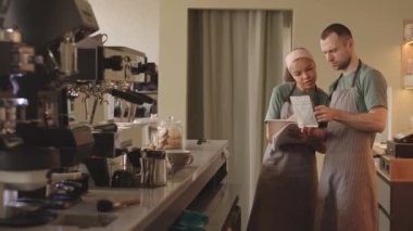Orta yaşlı beyaz erkek barista ve yeni melez kız arkadaşının kafede durup kahve paketini tartışırken orta boy bir fotoğrafı. Not defterine not alan kadın