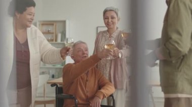 Mutlu Afrikalı kadın, tekerlekli sandalyedeki özürlü beyaz adam, gri saçlı Asyalı kadın ve dairede duran tanınmayan adam bardaklarını tokuşturup beyaz şarap içiyorlar.