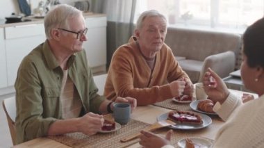Beyaz saçlı iki beyaz adamın yüksek açılı yakın plan fotoğrafları. Masalarında oturmuş tatlı pasta yiyor, tanınmayan insanlarla konuşuyorlar.