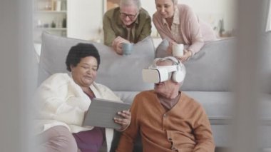 Orta halli, mutlu, kıdemli arkadaşların evdeki koltukta oturduğu, VR kulaklık takan bir adam, tablet bilgisayarı olan Afrikalı bir kadın, ellerinde fincanlarla onlara bakan gri saçlı bir çift.
