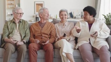 Orta uzunlukta, çok ırklı dört kıdemli arkadaş oturma odasında kanepede oturup, görünmez TV seyrediyor, parmaklarını ileri uzatıyorlar, konuşuyorlar ve gülüyorlar.