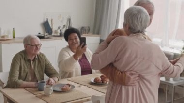 Orta boylu, beyaz saçlı, Asyalı bir kadının, kıdemli beyaz kocasıyla evde dans ederken, arkadaşlarının gülümserken, masada otururken, onları cep telefonuyla çekerken.