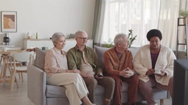 Oturma odasındaki kanepede oturan, televizyon seyreden, sohbet eden dört kıdemli arkadaşın orta uzunluğunda.