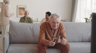 Orta boylu, beyaz bir adam oturma odasında kanepede oturuyor, duygusal olarak televizyon izliyor, arkadaşını çağırıyor, haberleri paylaşıyor.