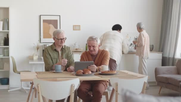 两个白种人的中年男人坐在房间的桌子旁 聊着天 与糕点一起喝茶 用平板电脑 女人在后台交流 — 图库视频影像