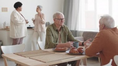 Orta uzunlukta beyaz, yaşlı bir adam masada oturuyor, arkadaşlarıyla çay içerken sohbet ediyor, çok ırklı kadınlar onlara katılıyor, arka plandan geliyorlar.