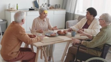 Orta uzunluktaki çok ırklı üst düzey arkadaşlar yemek masasında oturmuş, gülümsüyor, konuşuyor, Asyalı kadınların kazanmasını alkışlıyorlar.