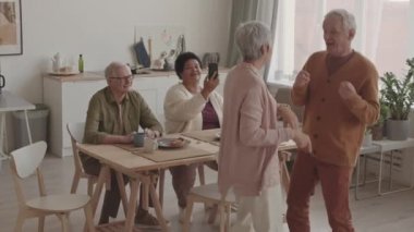 Yüksek açılı, orta boylu, gri saçlı, Asyalı bir kadın kıdemli beyaz kocasıyla dans ediyor. Çeşitli arkadaşları onları akıllı telefondan yemek masasında otururken filme çekiyor.