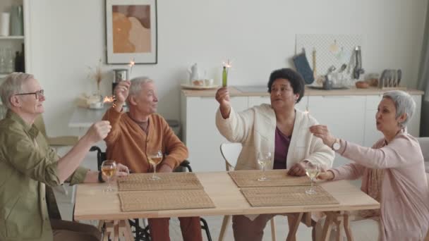 中等身材的亚洲和非洲妇女 老年残疾人和白人男子坐在餐厅的餐桌前 手持点燃的孟加拉灯微笑着 — 图库视频影像