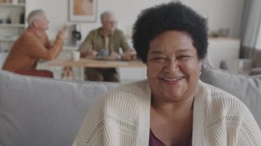 Orta yaşlı mutlu Afrikalı bir kadının göğüs göğüse duruşu. Koltukta oturuyor, yukarı bakıyor ve kameraya gülümsüyor.