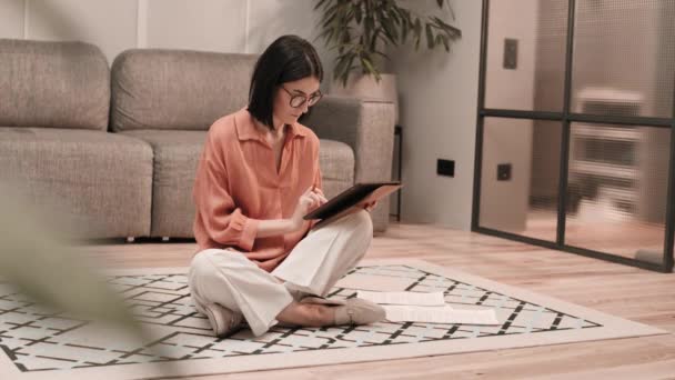 照片中年轻聪明的白人女人戴着眼镜 双腿交叉坐在公寓的地板上 用平板电脑翻阅文件 — 图库视频影像