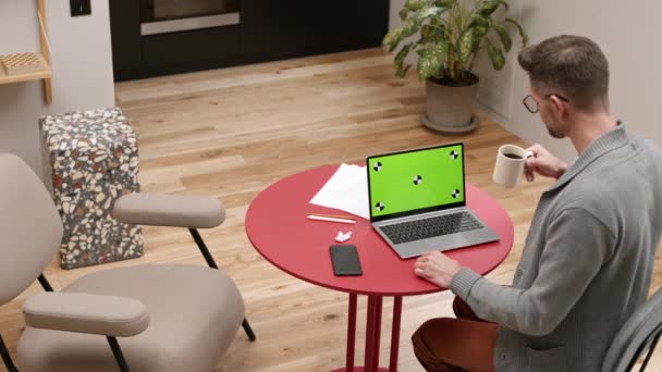 深色头发男子的高角形后背坐在红色桌旁 开着绿色屏幕的笔记本电脑 在家里喝咖啡 — 图库视频影像
