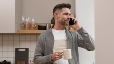 Beyaz kupa tutan, gülümseyen, cep telefonuyla konuşan, mutfakta bekleyen, telefonu kapatan neşeli beyaz bir adam.