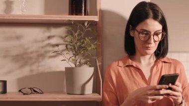 Koyu renk saçlı, beyaz bir kadının göğsü. Elinde bitki saksısı ve nesnelerle rafların yanında duruyor. Cep telefonu kullanıyor.