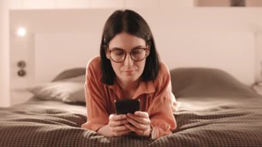 Beyaz tenli, gözlüklü, şeftali rengi gömlekli, evde yatağın üstüne uzanmış, akıllı telefon kullanan,