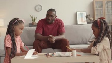 Orta uzunluktaki Afrikalı genç bir adam oturma odasında oturmuş kahve masasında resim çizen iki küçük sevimli kızı seyrediyor ve onunla konuşuyor.