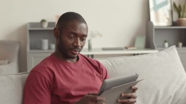 Koltukta oturan, tablet bilgisayar kullanan neşeli, genç Afrikalı adamın orta boy yakın çekimi. Şirin kızları ona hediyelerle sürpriz yapıyor. Kanepede zıplıyor.