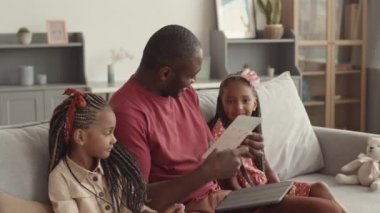 Orta uzunluktaki neşeli Afrikalı baba oturma odasında oturuyor, sevimli küçük kızlarına sarılıyor, gülümsüyor.