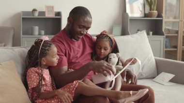 Orta uzunlukta neşeli Afrikalı bir adam kanepeye oturmuş gülümsüyor, hediyelerini açıyor, tatlı kızlarına sarılıyor. 