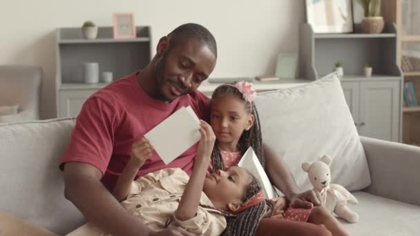 中年快乐的年轻非洲父亲坐在客厅的沙发上 抱着他可爱的小女儿 看着手工制作的明信片 — 图库视频影像