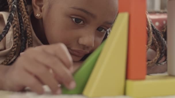 床に横たわる愛らしい小さなアフリカの女の子のクローズアップ カラフルな建物のおもちゃの作品で遊んで それを見て楽しむ — ストック動画