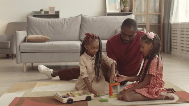 広いショットの若い幸せなアフリカの父と彼の愛らしい小さな娘のリビングルームの床に座って カラフルな木製のおもちゃのレンガで遊んで — ストック動画