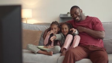 Genç, neşeli Afrikalı bir adam ve iki sevimli kız kanepede oturmuş televizyon izliyor ve gülümsüyor.