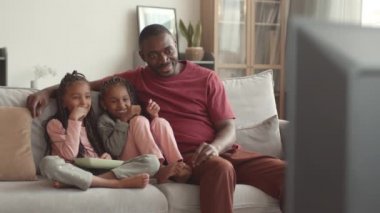 Genç ve mutlu Afrikalı bir adam ve iki sevimli kızın evdeki kanepede oturup televizyon izlemesi.