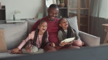 Yüksek açılı genç Afrikalı bir adam ve iki sevimli kadın çocuk evde kanepede oturmuş televizyon izliyor, gülüyor, abur cubur yiyorlar.