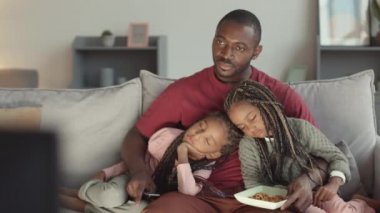 Orta uzunlukta, kanepede oturup televizyon izleyen, iki kolunda da örgü takan sevimli küçük kızları olan yorgun Afrikalı bir adam.