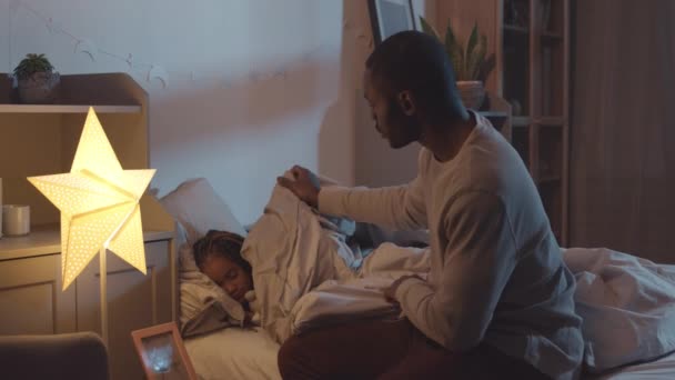 侧视图中等长度的年轻非洲父亲坐在孩子房间的床沿上 为他的小女儿铺上毯子 拍拍她的头 — 图库视频影像