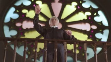 Beyaz yakalı, siyah elbiseli, beyaz yakalı beyaz bir papazın vitraylara karşı Hıristiyan kilisesinde durması ve Pazar vaazları vermesi.