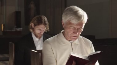 Lutheran kilisesindeki ahşap banklarda oturan ve Pazar ayini sırasında İncil okuyan farklı yaşlarda iki beyaz adamın orta boy fotoğrafı.