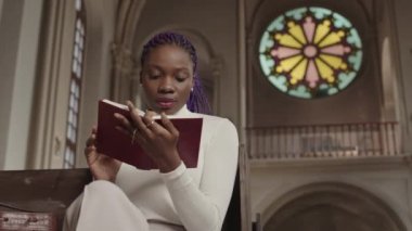 Lüteriyen kilisesinde ahşap sıralarda oturan ve İncil okuyan mor saçlı Afrikalı genç bir kadının orta boy portresi.