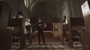 Lüteriyen kilisesinde elinde kutsal İncil 'le dikilen siyah giysiler, maske ve beyaz yaka giyen ve cemaatin önünde maskeli bir şekilde konuşan Afro-Amerikan din adamını yakınlaştır.