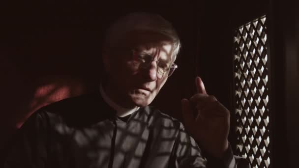 黒い衣 白い襟と眼鏡を手に聖書と一致し 木製の窓を介して教区に話を座っている古い白人の司祭の中のショット — ストック動画