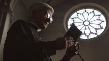 Beyaz yakalı, siyah cüppeli, beyaz yakalı yaşlı beyaz papazın elinde kutsal İncil ve tespih boncuklarıyla Lüteriyen kilisesinde durup dua edişinin düşük açılı görüntüsü.