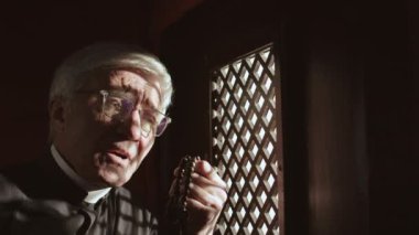 Kafkas rahip cüppeli, beyaz yakalı ve gözlüklü, elinde tespih boncuklarıyla günah çıkarma kabininde oturan kilise cemaatini dinleyen ve sonra haç çıkartan