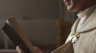 Hristiyan kilisesinde elinde kutsal kitapla dikilen ve kitap okuyan, tanınmamış, yaşlı, şenlikli, altın bir cübbe giyen rahibi yakından takip edin.