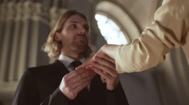 Düğme delikli siyah takım elbiseli beyaz damadın kilisedeki düğün töreninde tanınmayan gelininin parmağına taktığı düşük açı.