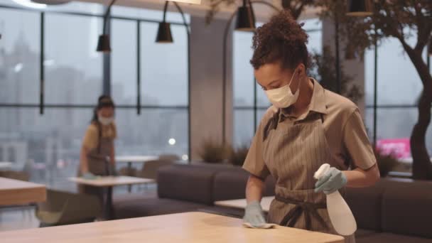 顔のマスクと手袋を着用し テーブルの上に液体を消毒噴霧し ラグでそれを拭き取る若い混合レースの女性カフェ労働者の中程度のショット — ストック動画