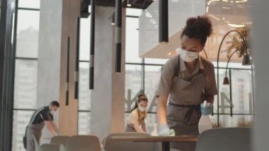 Yüz maskesi ve eldiven takmış, modern restoranda dezenfekte sıvısı ile masaları silen, arka plan üzerinde çalışan genç, melez kafe çalışanlarının düşük açılı görüntüsü.