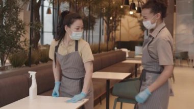 Orta uzunlukta bir Asyalı garson, dezenfektanla kahve masasını temizliyor, kadın melez meslektaşı geliyor ve onu dirsek seğiyle selamlıyor, maskeli ve eldivenli kadınlar, gülümsüyor ve konuşuyor.