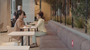Çok ırklı iki genç bayan arkadaşın gülümsemesi, konuşması, restoranda masada oturması, eldivenli erkek garson ve yüz koruyucusu geliyor.