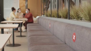 Koltuğun arkasında haç işaretleri olan kırmızı uyarı levhaları olan geniş bir kanepe. Maskeli üç farklı arkadaş restoranda masada oturuyor.